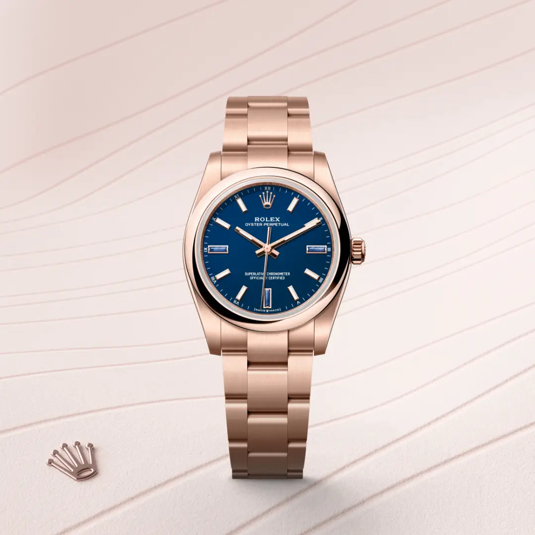 m124205-0002-slide1 | Brela Rose gold Rolex Oyster Perpetual watch with a blue dial on a pale striped surface, with the Rolex crown logo nearby. | Brela