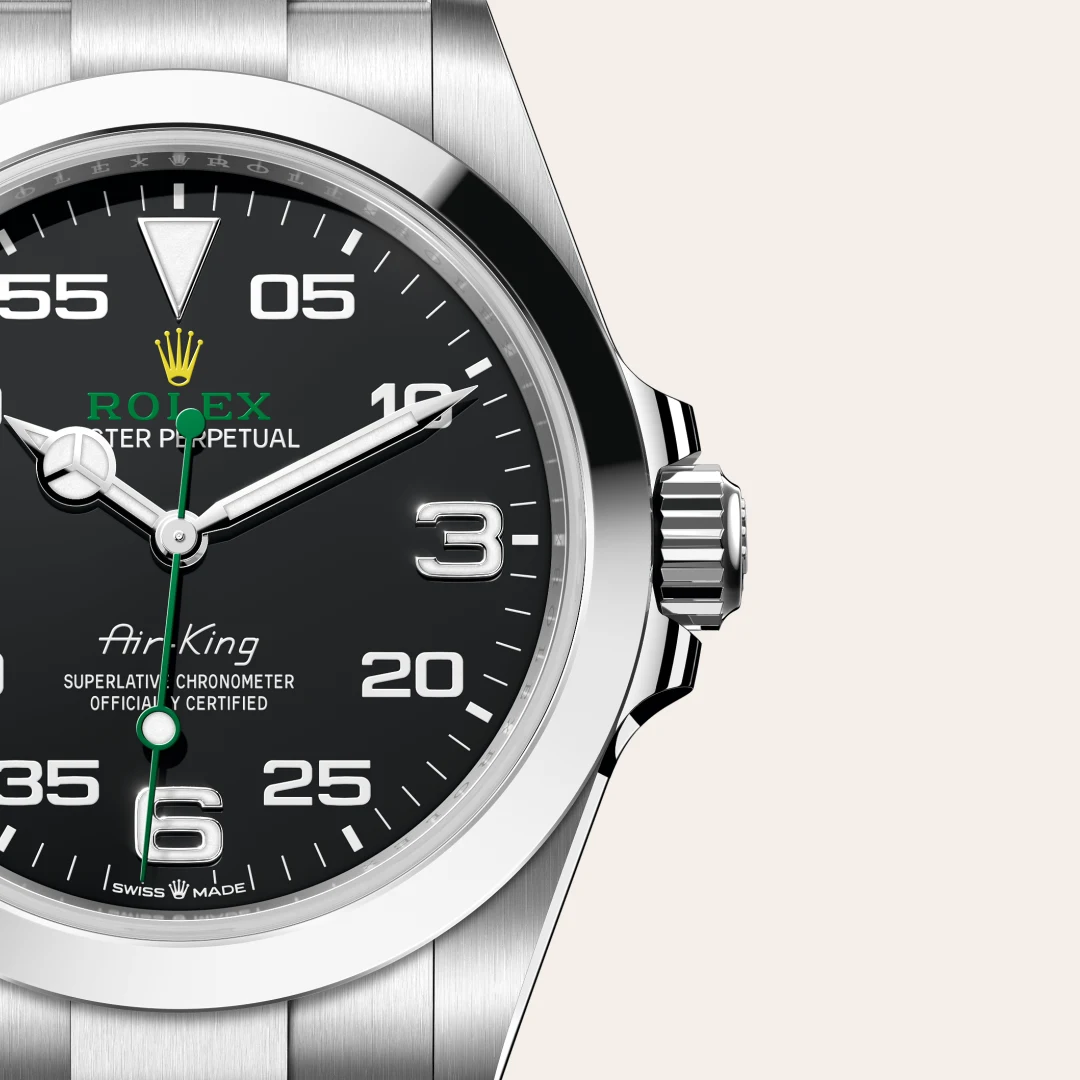 Close-up of a Rolex Air-King watch face with a black dial, green logo text, gold crown, and silver hands on a stainless steel case. | Brela