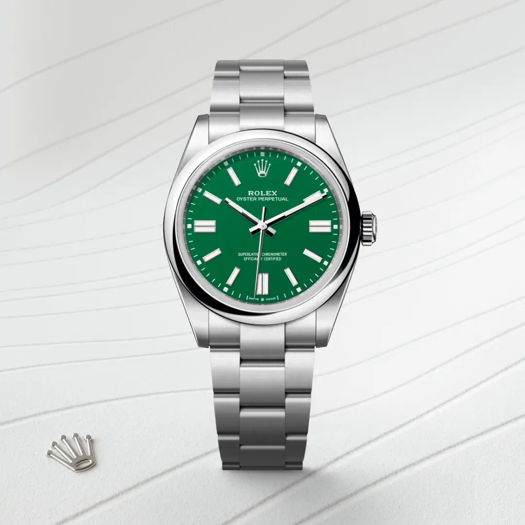 m134300-0004-slide1 | Brela Rolex Oyster Perpetual watch with a green dial and stainless steel bracelet, shown with a small Rolex crown emblem nearby on a light striped surface. | Brela