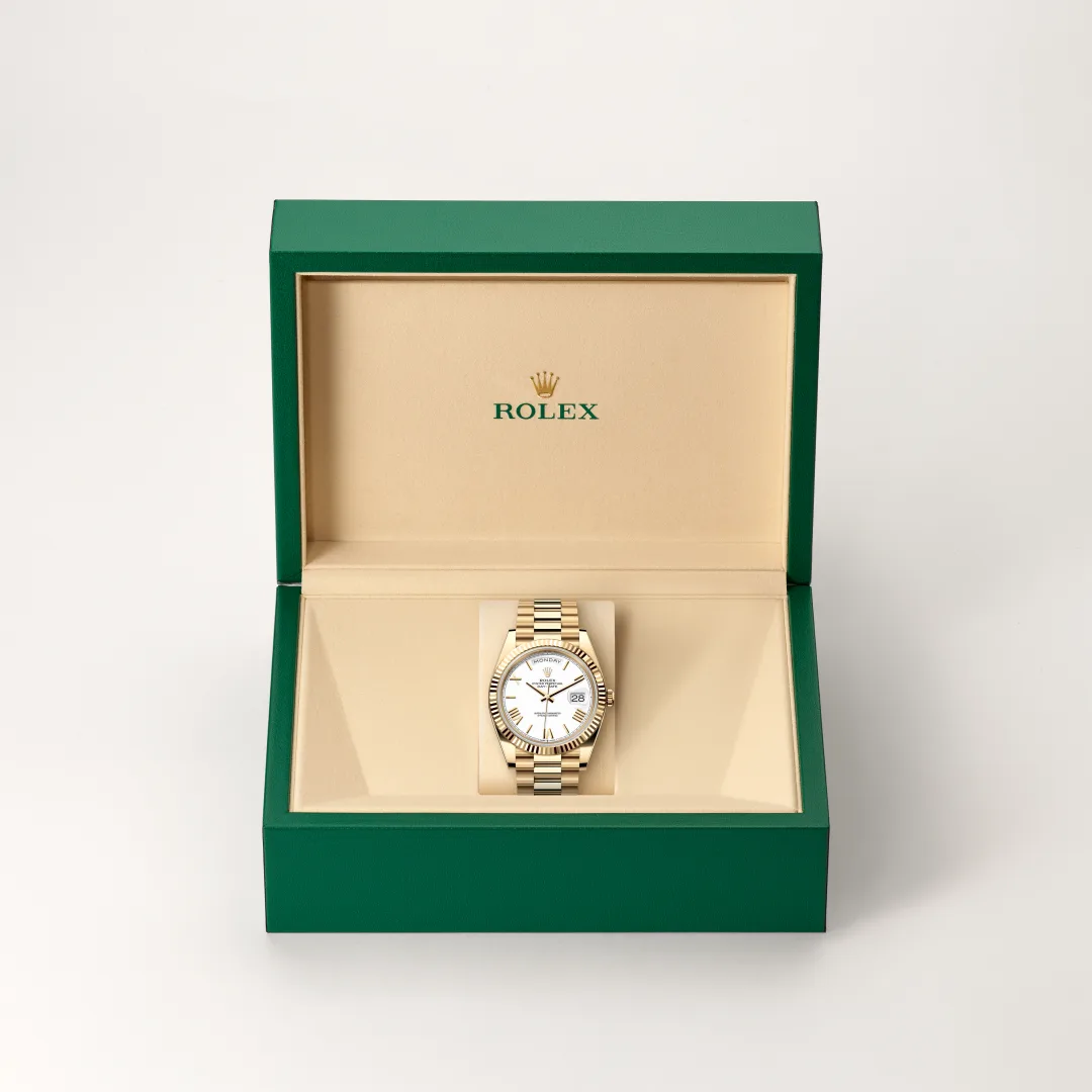 Gold Rolex watch in an open green presentation box with beige interior and the date window visible on the white dial. | Brela