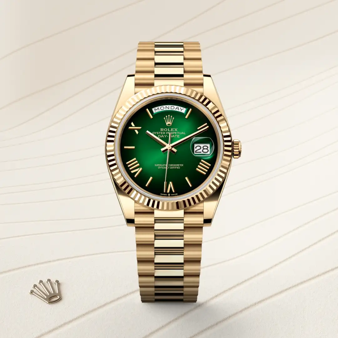 Gold Rolex Day-Date wristwatch with a green dial and date window on a pale surface; a small Rolex crown charm sits nearby. | Brela