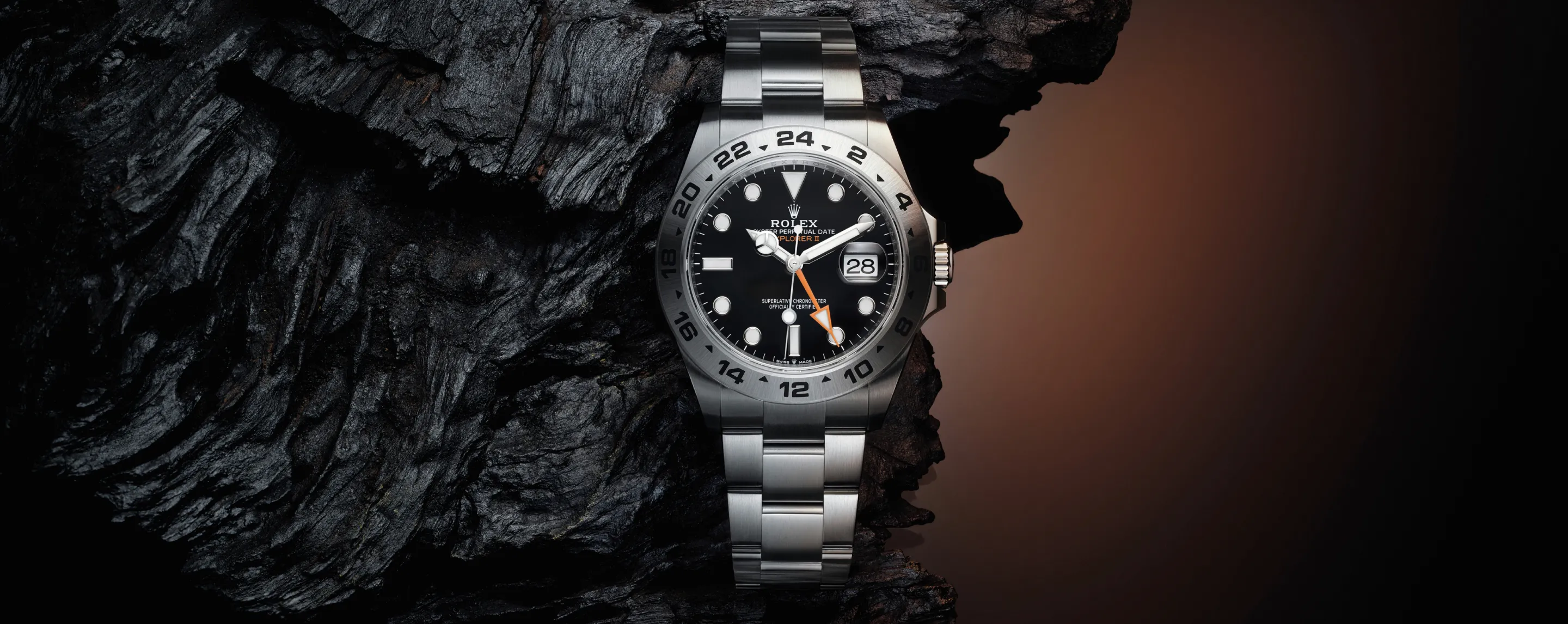 Rolex Explorer II stainless steel watch on dark rock, showing date at 3 o'clock and orange 24-hour hand.
