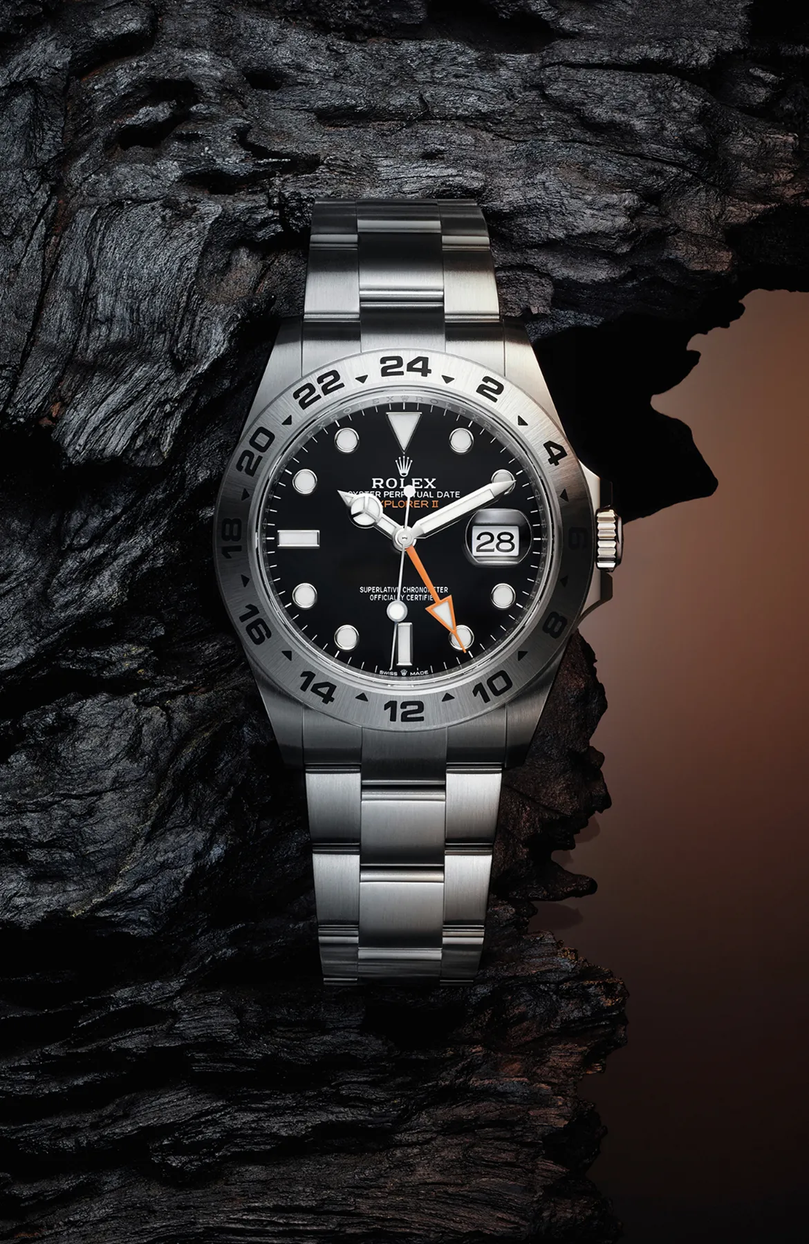 Close-up of a stainless steel Rolex watch with a black dial and orange 24-hour hand resting on rugged black rock surface.