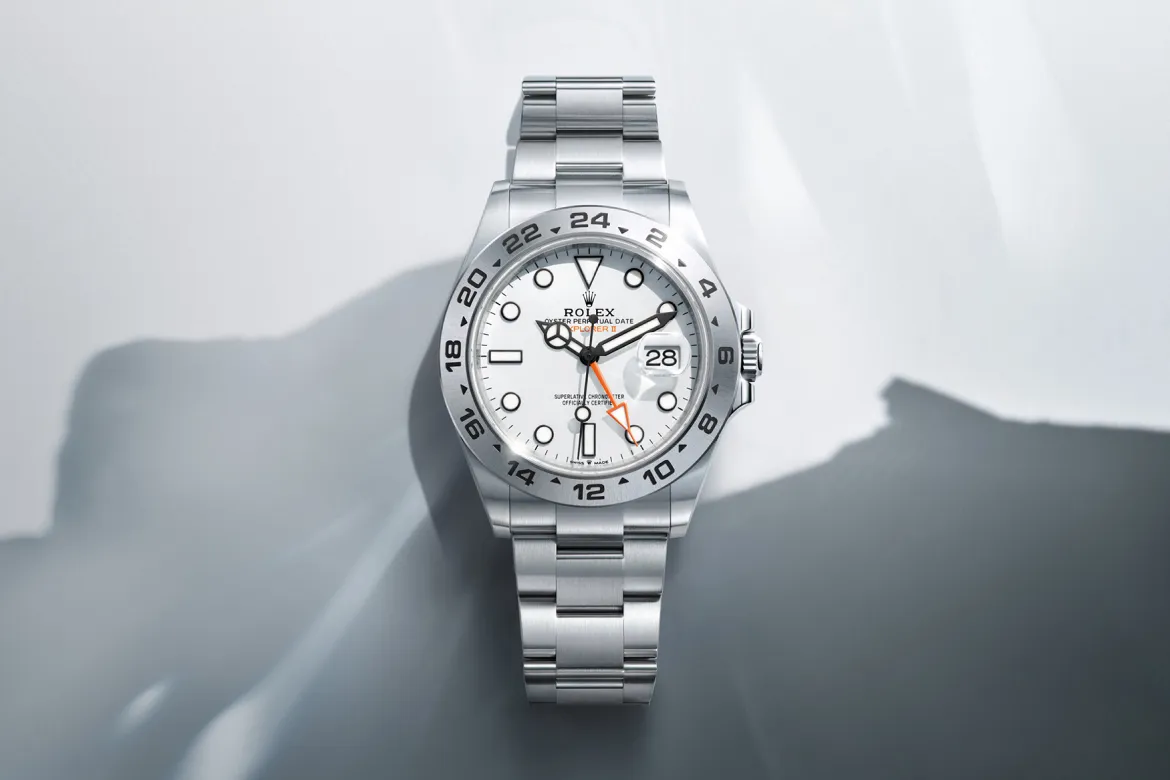 Stainless steel Rolex watch with white dial, date window at 3, and orange 24-hour hand on a pale background.