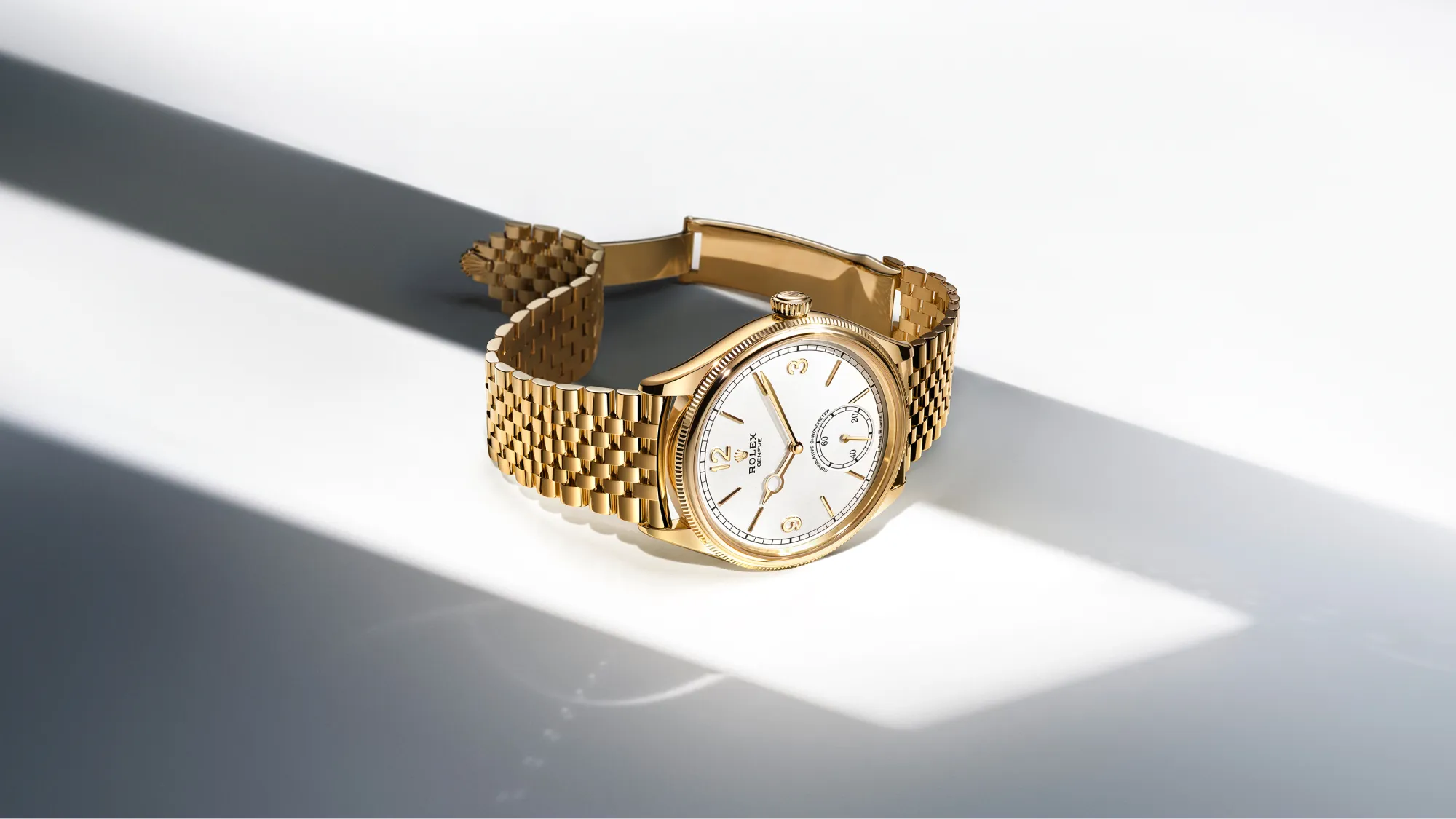 rolex-oyster-perpetual-universal-classic-style-m52508-0008-landscape Gold wristwatch with a white dial and gold bracelet resting on a white surface, diagonal shadows in the background.