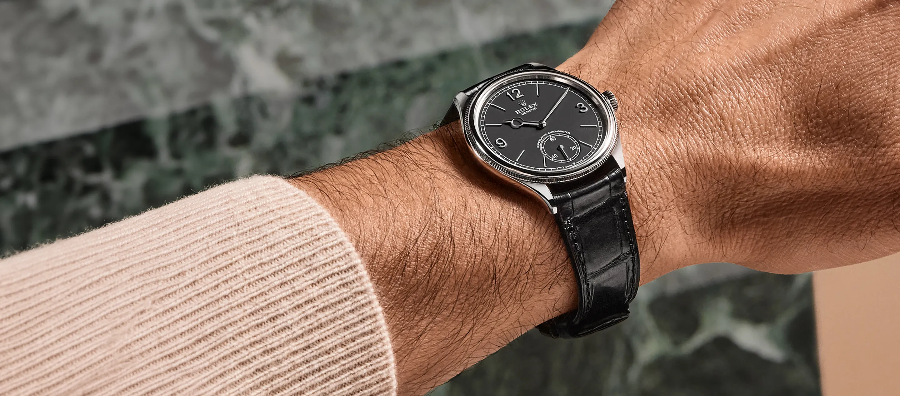 rolex-perpetual-1908-ambiance-m52509-0002-landscape Close-up of a black-dial Rolex wristwatch with a silver case and black leather strap on a hairy forearm wearing a beige knit sleeve