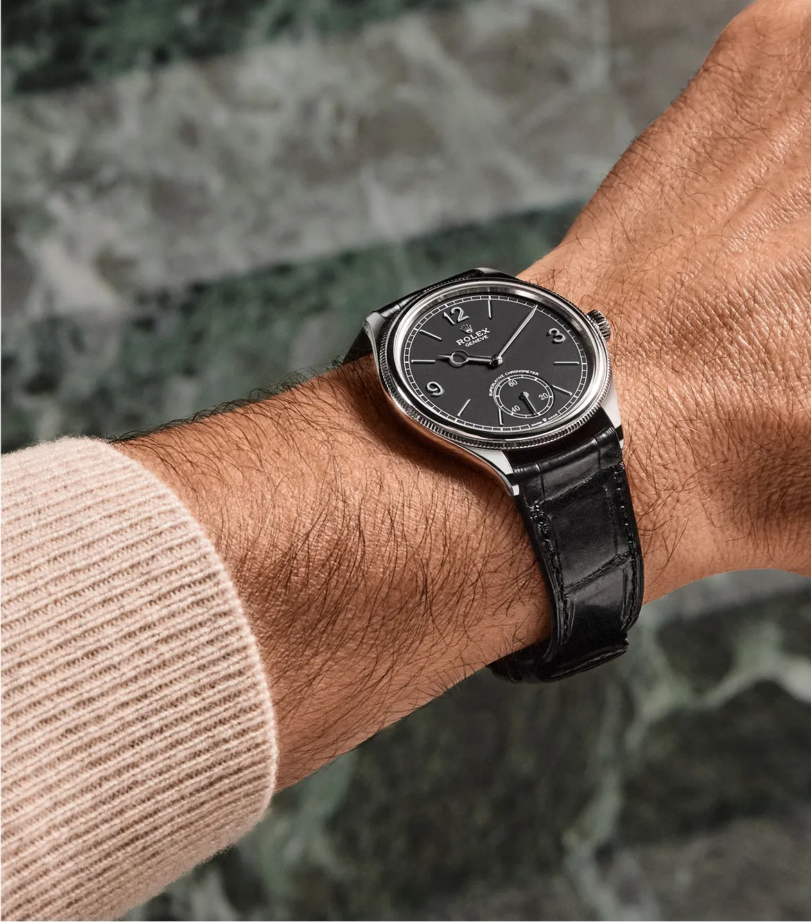 rolex-perpetual-1908-ambiance-m52509-0002-portrait Black dial Rolex wristwatch with silver case and black leather strap worn on a hairy forearm, beige knit sleeve visible in the corner.