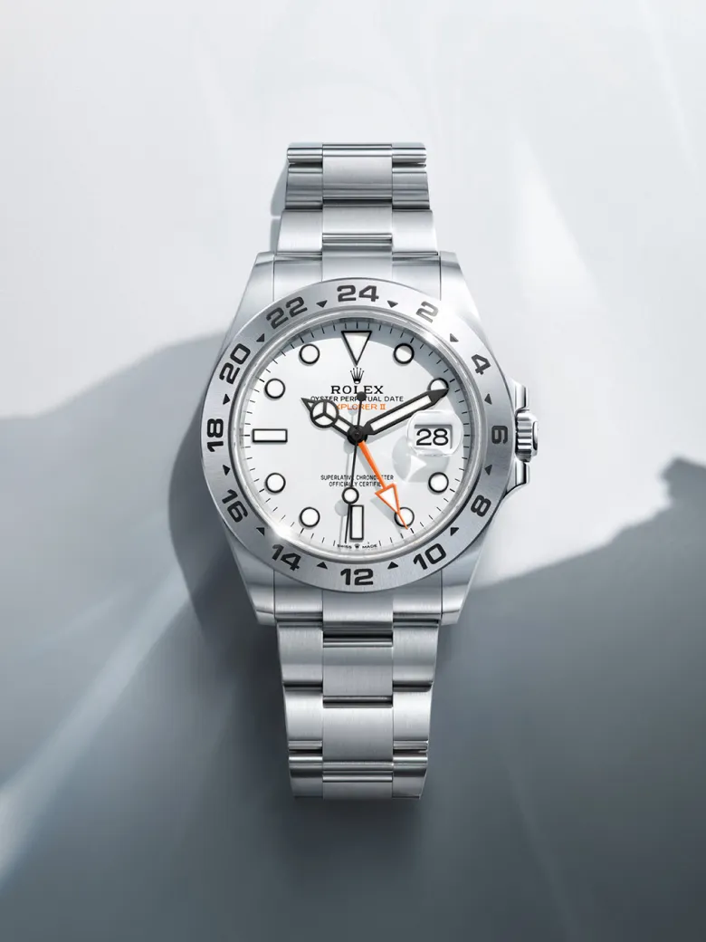 rolex-watches-explorer-ii-m226570-0001-portrait | Brela Close-up of a stainless steel Rolex Explorer II watch with a white dial, 24-hour bezel, and date window at 3 o'clock set against a soft gray background. | Brela