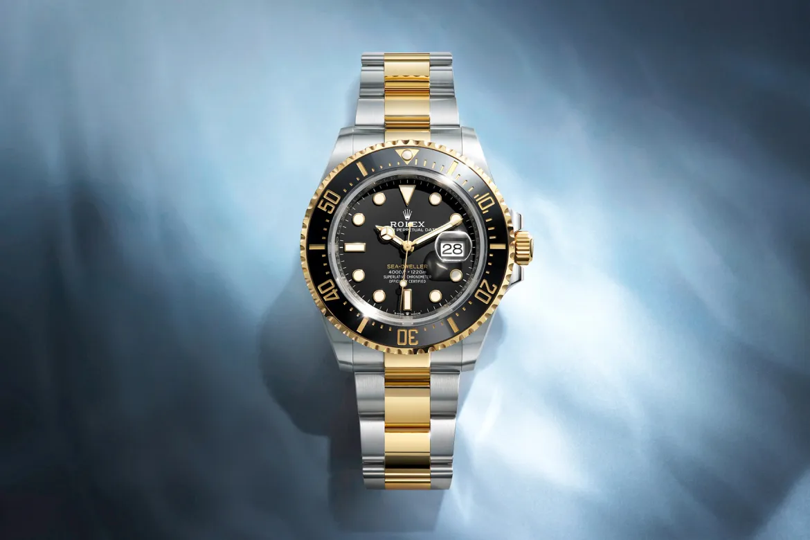 rolex-watches-sea-dweller-m126603-0001-landscape | Brela Two-tone Rolex watch with gold and silver bracelet, black bezel and dial, and date window, displayed on a blue gradient background. | Brela