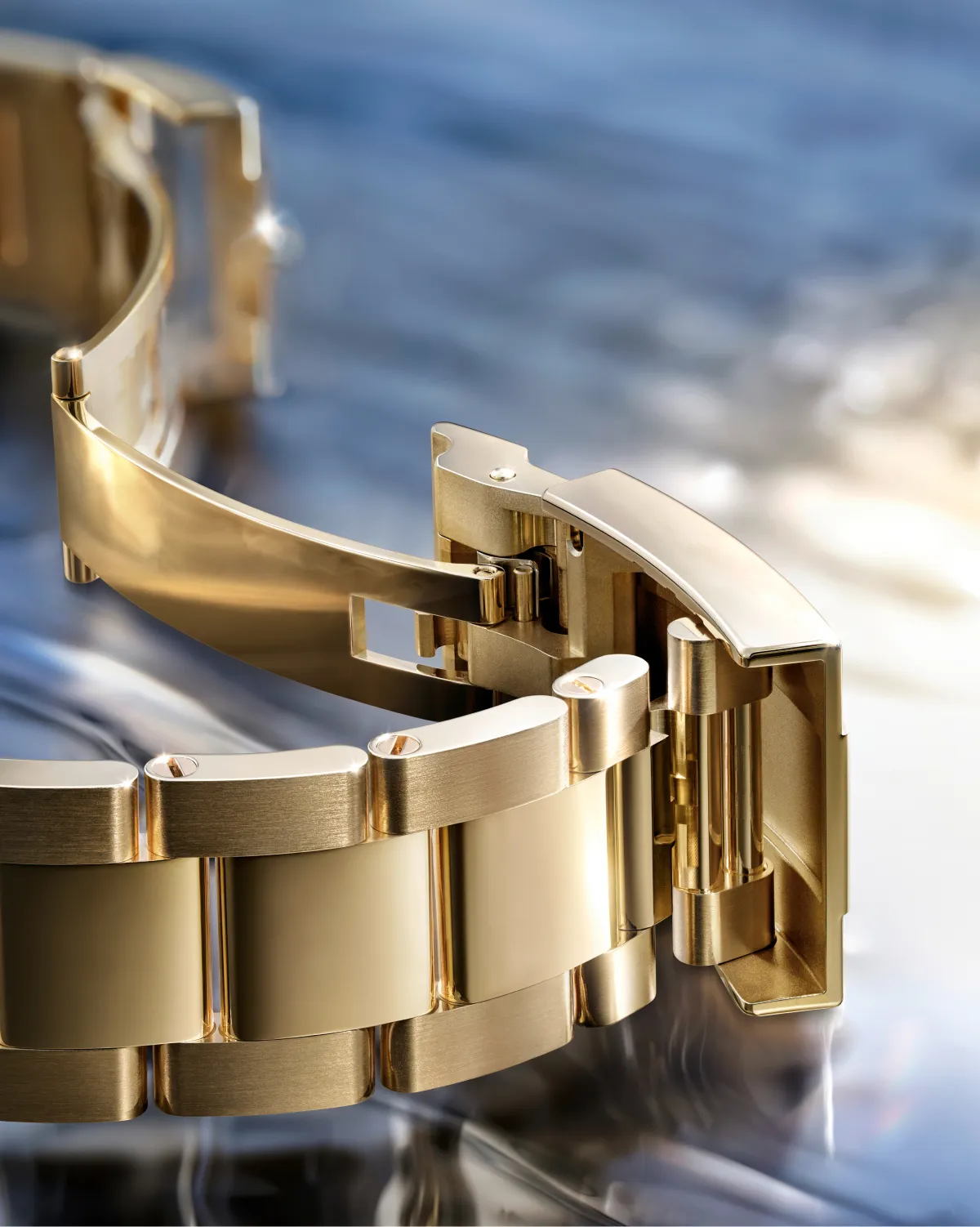 Close-up of a gold metal watch bracelet with a deployant clasp on a reflective surface.