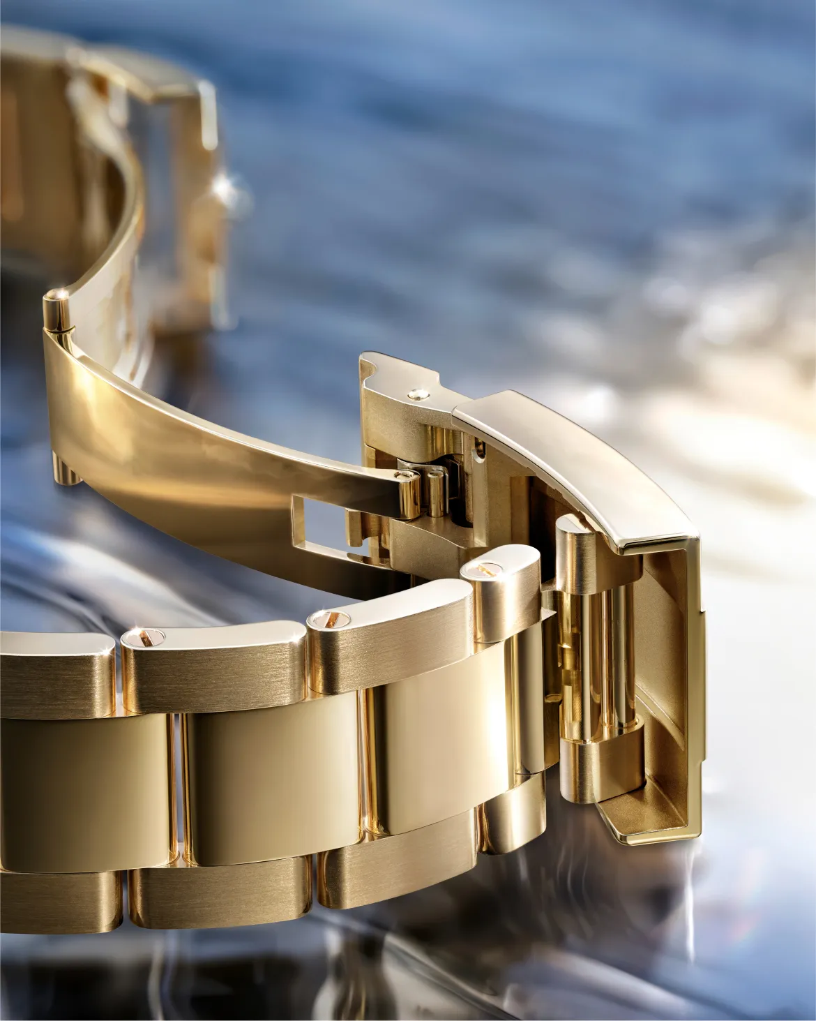 Close-up of a gold metal watch band with a deployment clasp on a reflective surface