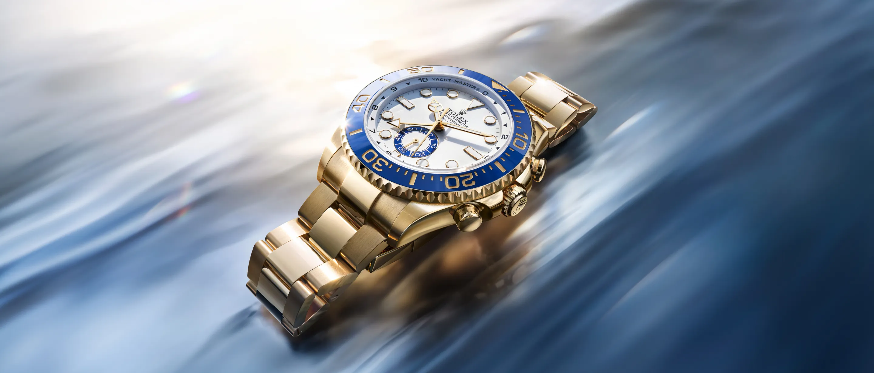 Gold Rolex wristwatch with a blue bezel resting on a blurred blue-gray background, angled to show its face and bracelet.