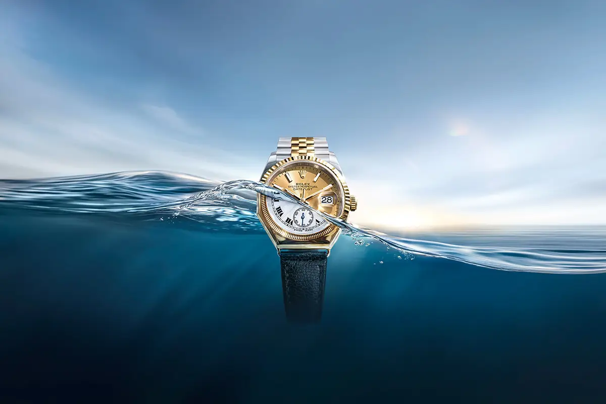 discover-rolex-oyster-story-m126233-0015_first_oyster_octagonal_1926_2604_0001-landscape | Brela Gold and silver Rolex watch partially submerged in blue water, with waves splashing around the face and date window visible. | Brela