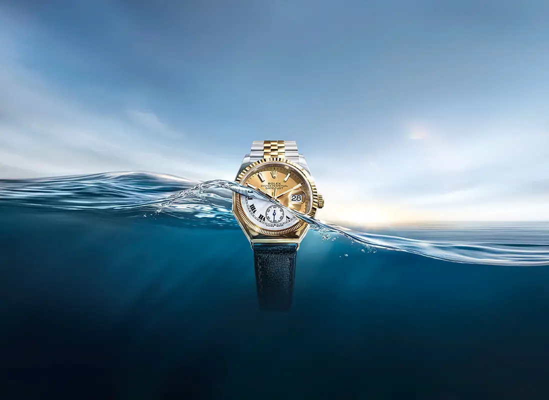 discover-rolex-oyster-story-m126233-0015_first_oyster_octagonal_1926_2604_0001-portrait | Brela Gold Rolex wristwatch with two-tone bracelet partially submerged in blue ocean, sunset on the horizon behind it. | Brela