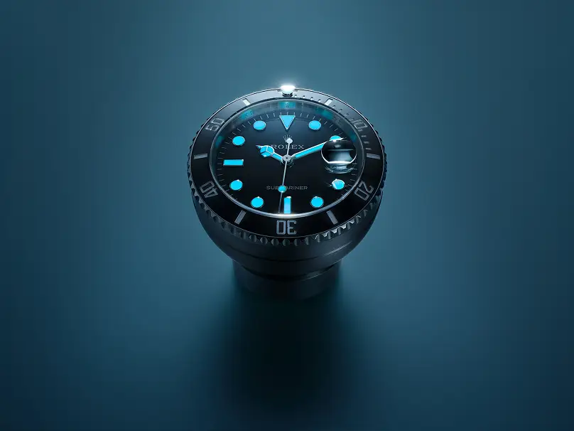 rolex-accessories-table-clock-night-c909010ln-0001_2512stj_002_srgb-landscape | Brela Close-up of a Rolex Submariner dive watch with black dial, blue luminous markers, and a magnified date window on a teal background. | Brela
