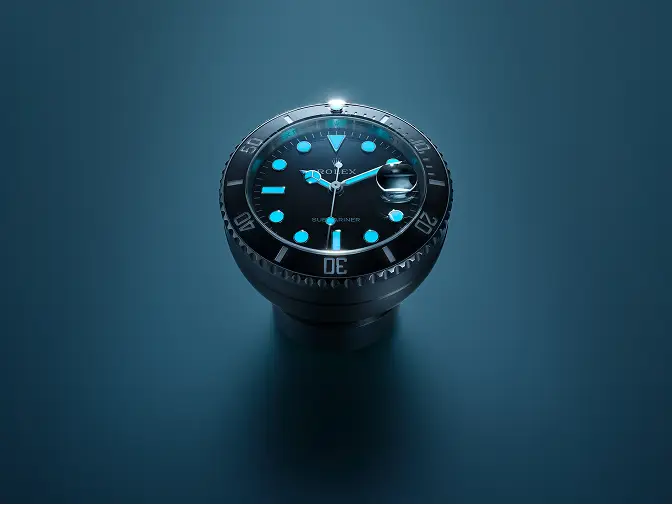 rolex-accessories-table-clock-night-c909010ln-0001_2512stj_002_srgb-portrait | Brela Close-up of a black Rolex Submariner watch with blue luminescent markers on a blue surface. | Brela