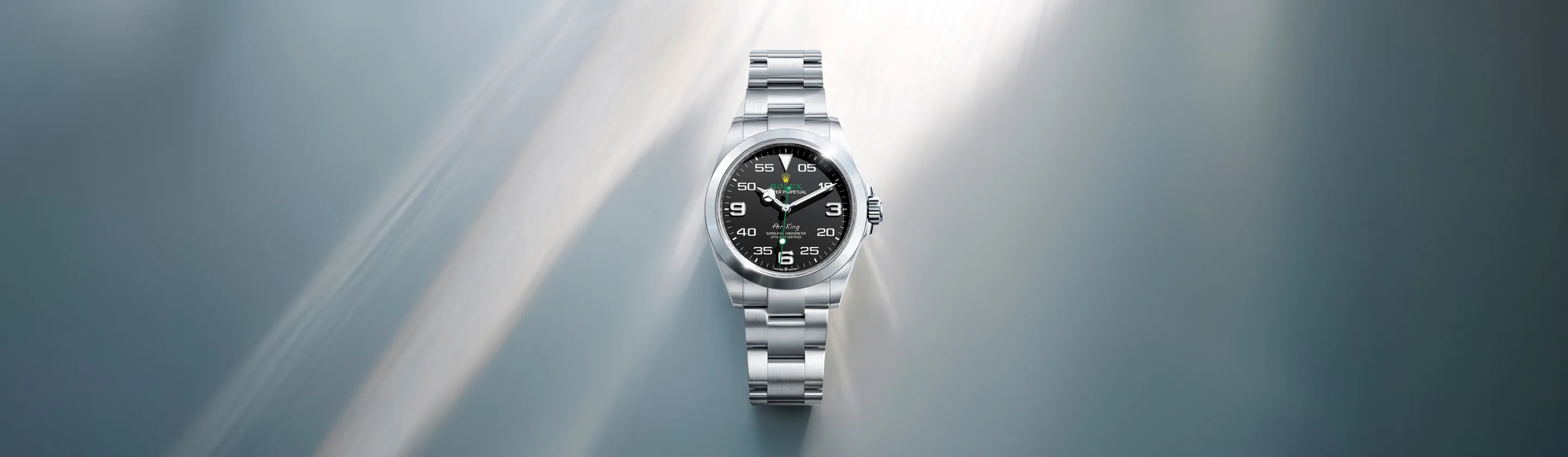 rolex-air-king-cover-m126900-0001-landscape Silver Rolex wristwatch with black dial and metal bracelet centered on a soft blue background.