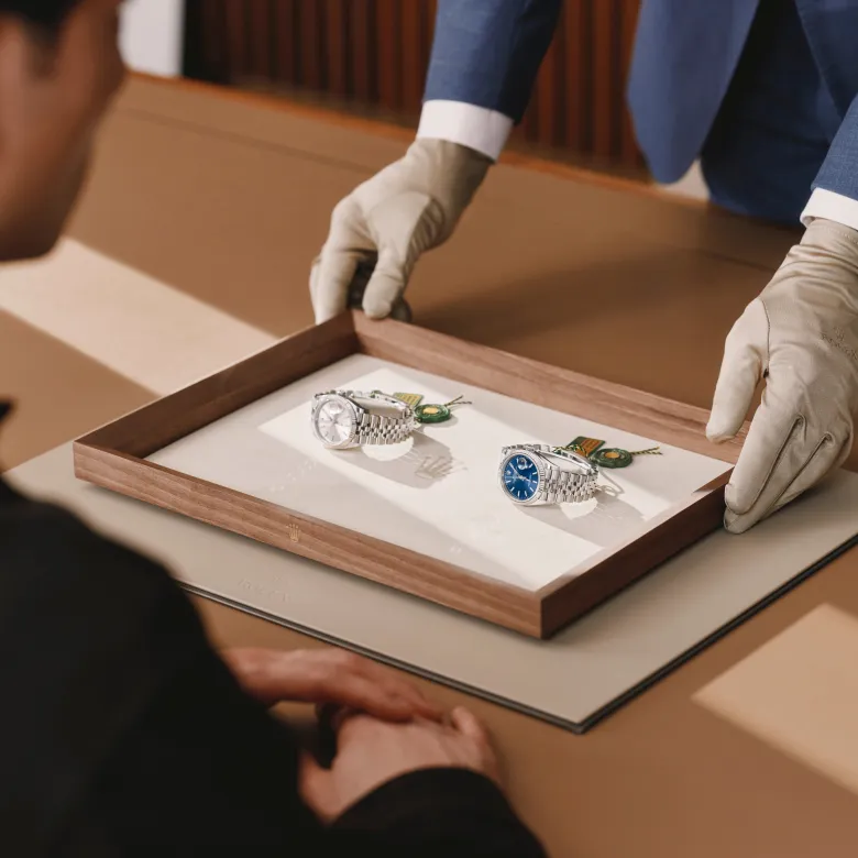 rolex-contact-message-elmt_2603fg_041-portrait | Brela Jeweler wearing gloves presents two wristwatches in a display tray to a customer seated at a counter. | Brela