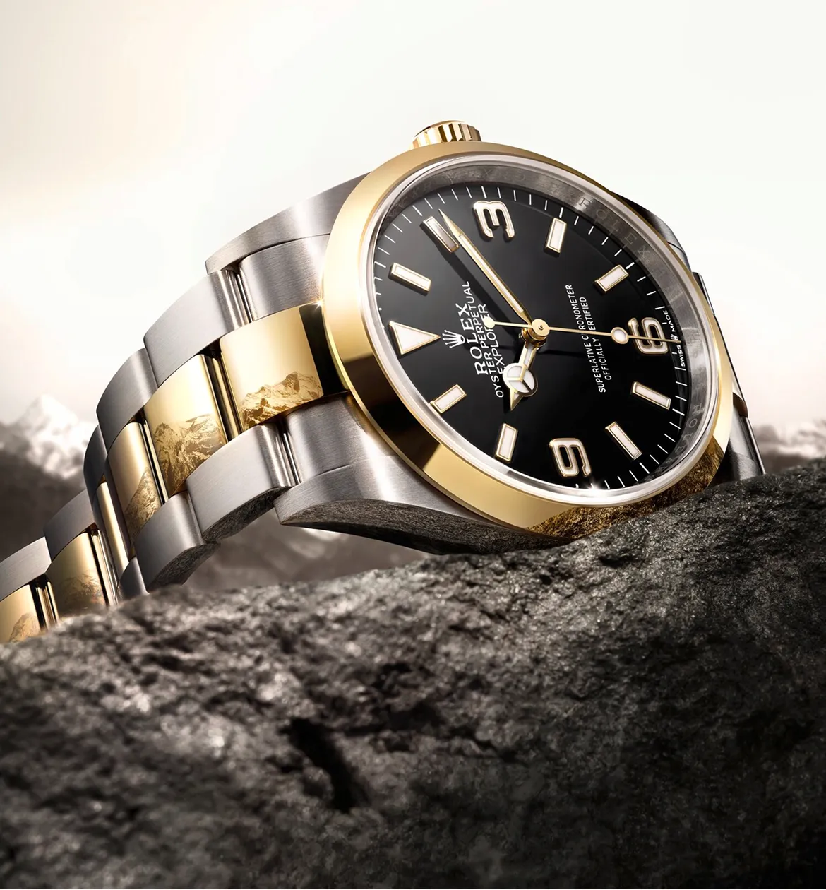 rolex-explorer-ambiance-m124273-0001-portrait Two-tone Rolex wristwatch with a black dial resting on a rocky surface, gold bezel and silver-gold bracelet.