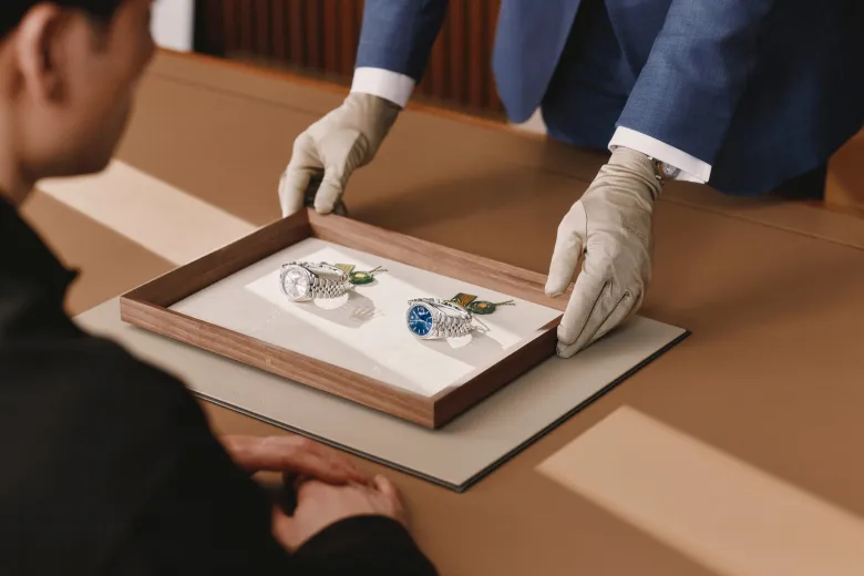 rolex-message-cover-portrait | Brela Two men wearing gloves present two luxury watches in a wooden display case for a client at a formal setting | Brela