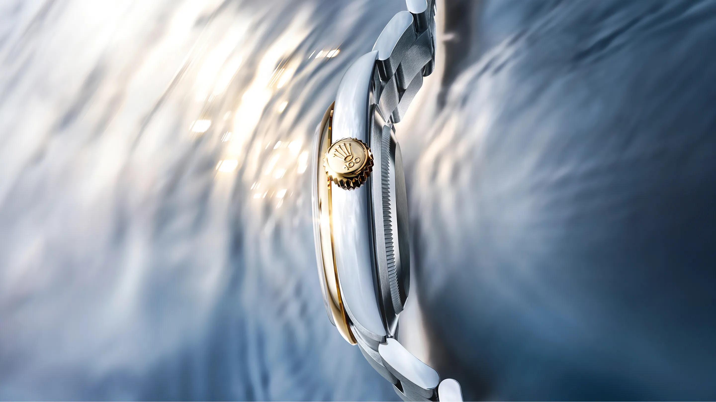 rolex-oyster-perpetual-100th-anniversary-ambiance-m134303-0001-landscape Close-up of a two-tone wristwatch crown and case with a gold crown and silver bezel against a blurred blue background, showing attention to detail.