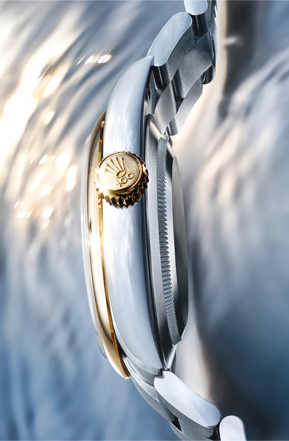 rolex-oyster-perpetual-100th-anniversary-ambiance-m134303-0001-portrait Close-up of a gold watch crown on a two-tone luxury wristwatch, diagonal angle showing the brushed steel case and crown detail