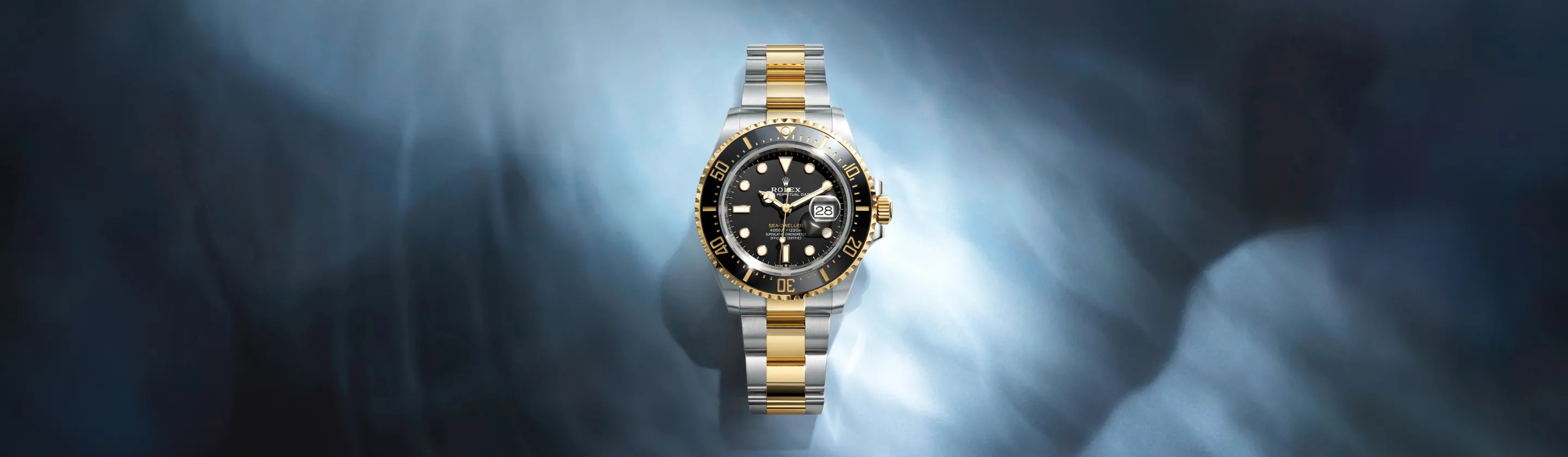 rolex-sea-dweller-cover-m126603-0001-landscape Two-tone Rolex watch with gold and silver bracelet, black dial, and date window, centered against a blue smoky background.