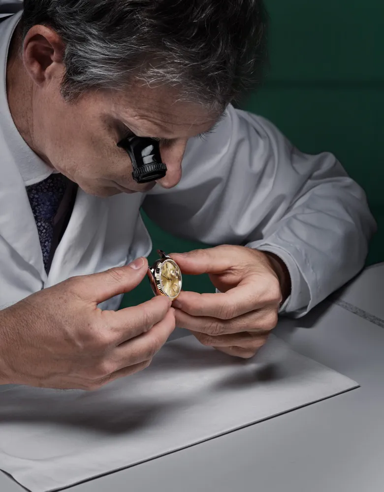 rolex-servicing-2234_rolex-sav_retailers_104_estimation_vérification_no_de_serie_boite_v5-portrait | Brela Watchmaker wearing a loupe and white lab coat examining a gold wristwatch at a workbench, close-up | Brela