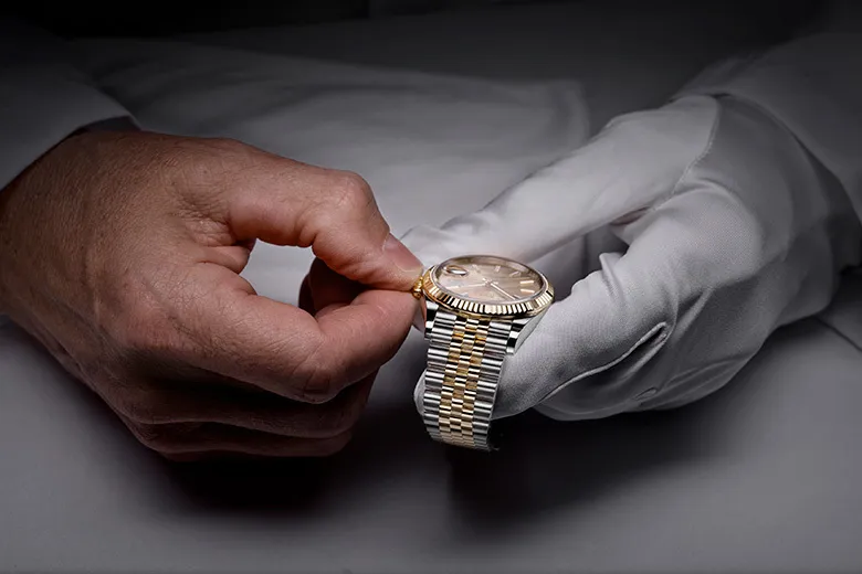rolex-servicing-cover_sav_1910dh_001-portrait | Brela Person wearing white gloves adjusts a two-tone gold and silver wristwatch on their wrist. | Brela