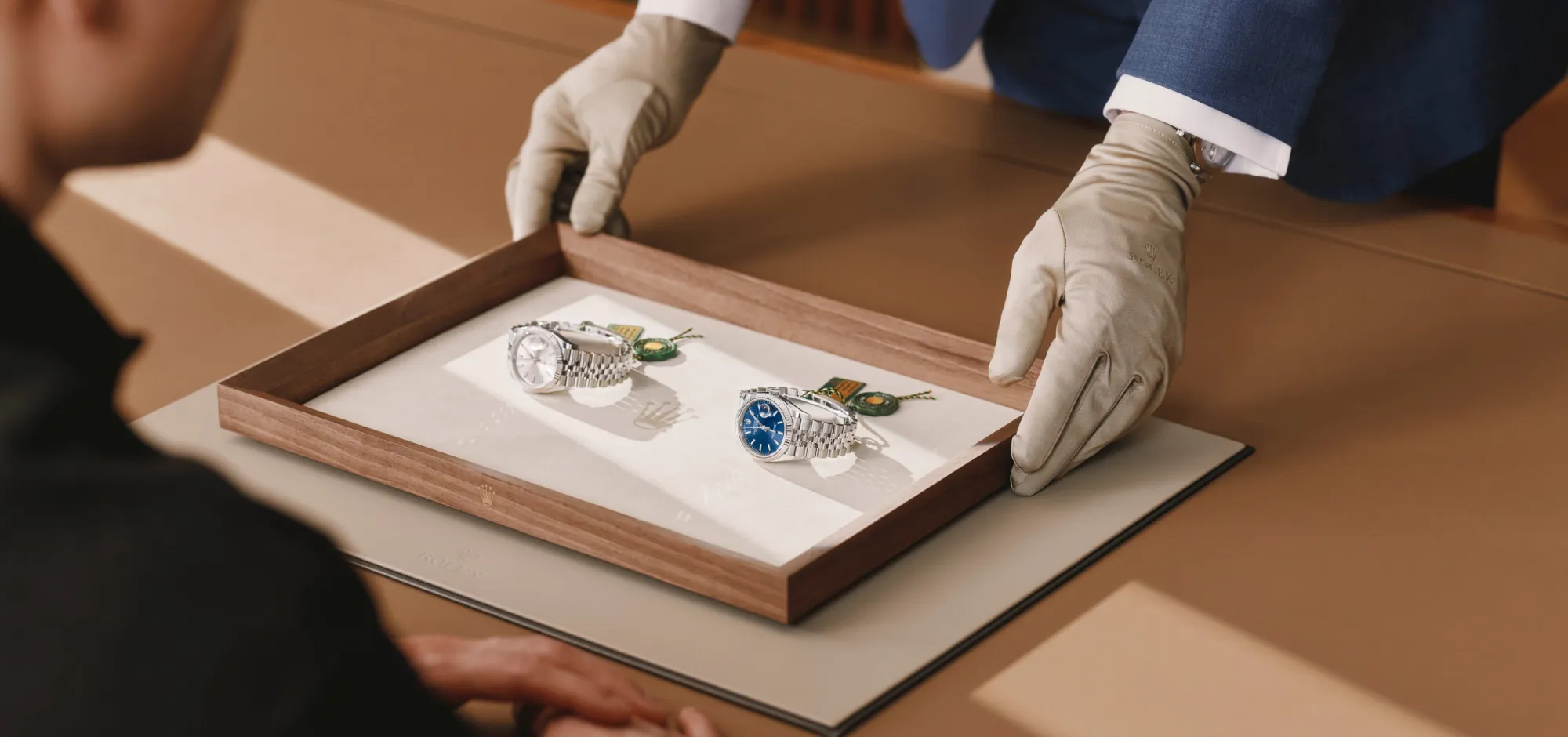 rolex-servicing-elmt_2603fg_041-landscape | Brela Two silver wristwatches in a wooden display box on a white mat, being adjusted with gloved hands by a person in a suit jacket: front and center blue-dial watch visible beside a second silver watch. | Brela