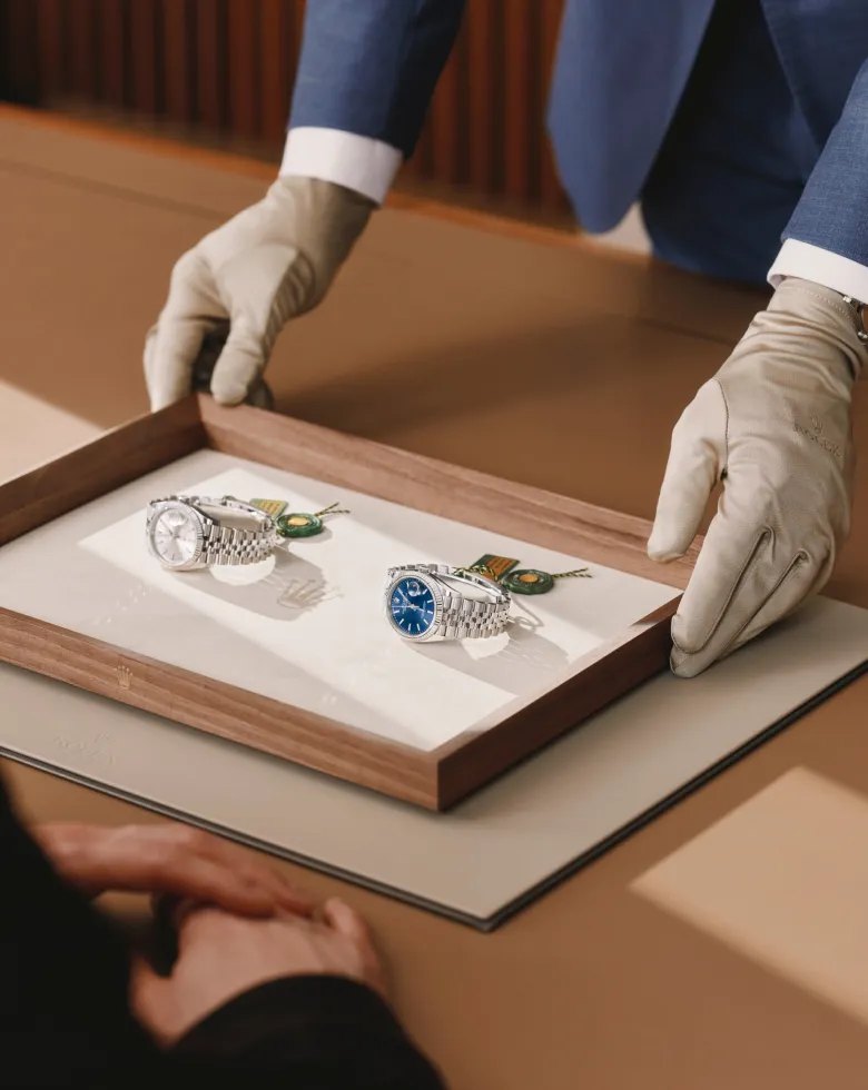 rolex-servicing-elmt_2603fg_041-portrait | Brela A jeweler wearing gloves presents two silver wristwatches on a wooden display tray to a customer. | Brela