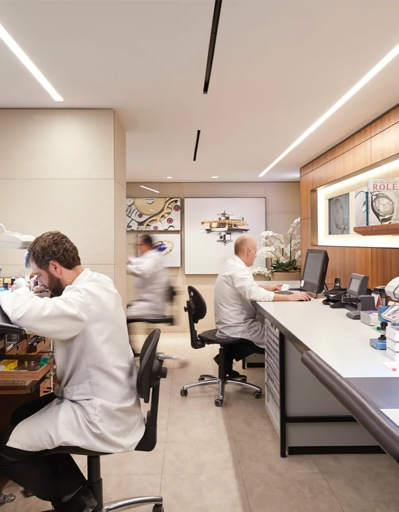 rolex-servicing-sav_barcelona_tousrolexboutique_2019gv_1124-portrait | Brela Watchmakers in white coats at workbenches in a modern repair workshop; one inspecting a piece with a loupe, others at computers along a counter. | Brela