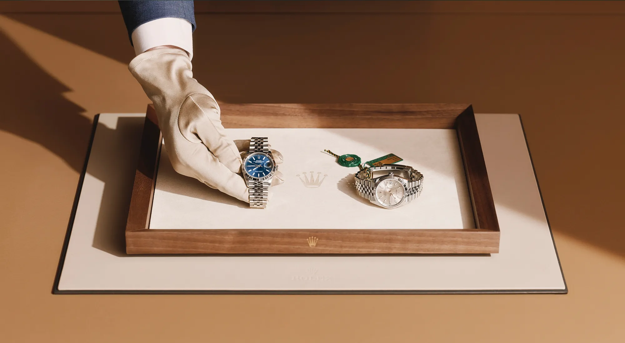 rolex-watchmaking-elmt_2603fg_074-landscape | Brela Gloved hand holding a silver Rolex with a blue face inside an open wooden display box, next to another silver watch with a metal bracelet. | Brela