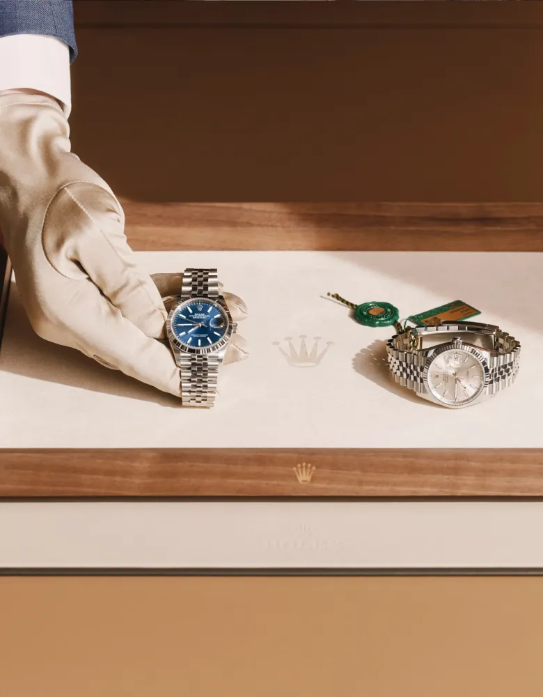 rolex-watchmaking-elmt_2603fg_074-portrait | Brela A gloved hand holds a blue-dial silver Rolex watch while another silver Rolex sits on the display tray beside it. | Brela