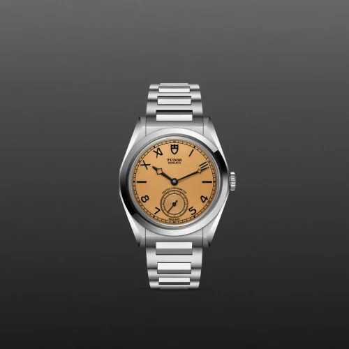 Stainless steel Tudor Monarch wristwatch with a tan dial on a dark gray background, shown from the front with the bracelet partially visible.