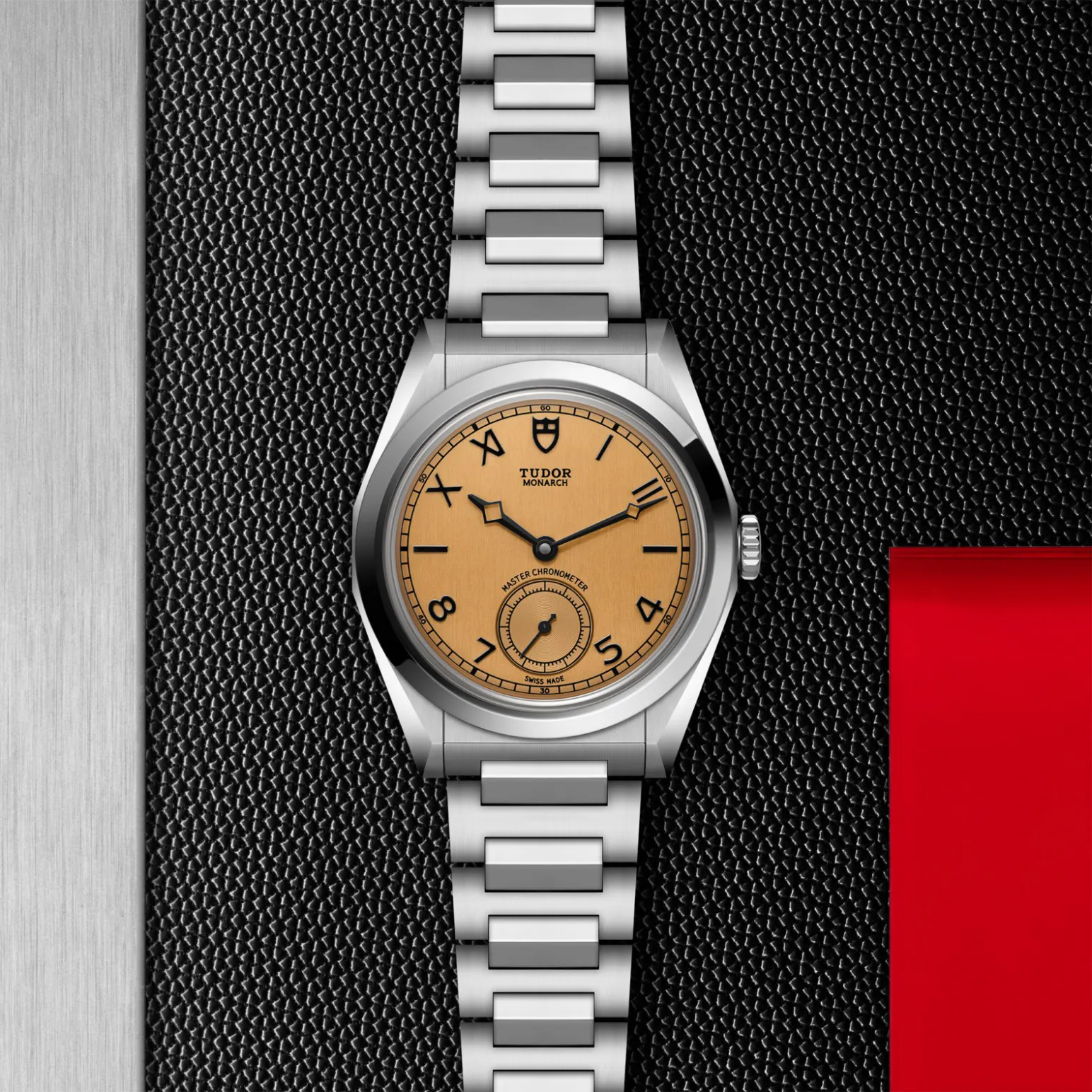 m2639w1a0u-0001in_store_flat_lay | Brela Stainless steel Tudor Monarch wristwatch with a tan dial on a textured black background and silver bracelet links | Brela
