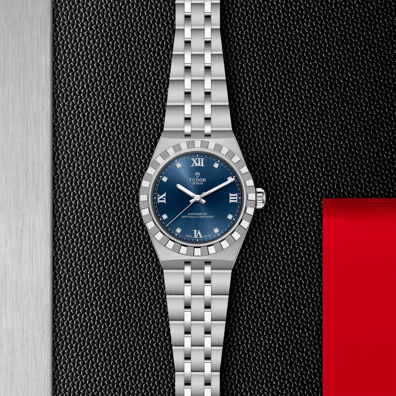 m2830a1a0-0001in_store_flat_lay | Brela Silver Tudor wristwatch with a blue dial, Roman numerals and diamond markers, centered on a textured black background. | Brela
