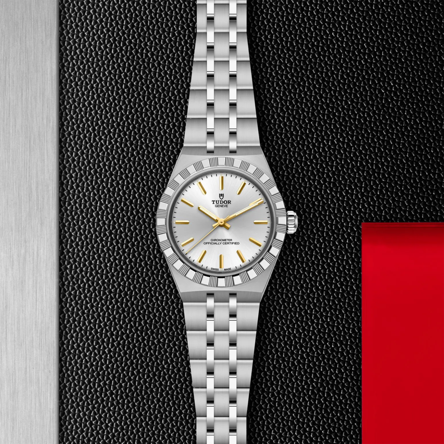 m2830a1a0-0002in_store_flat_lay | Brela Tudor stainless steel wristwatch with a silver dial and gold hour markers, centered on a black textured background with a silver left bar and a red panel on the right. | Brela