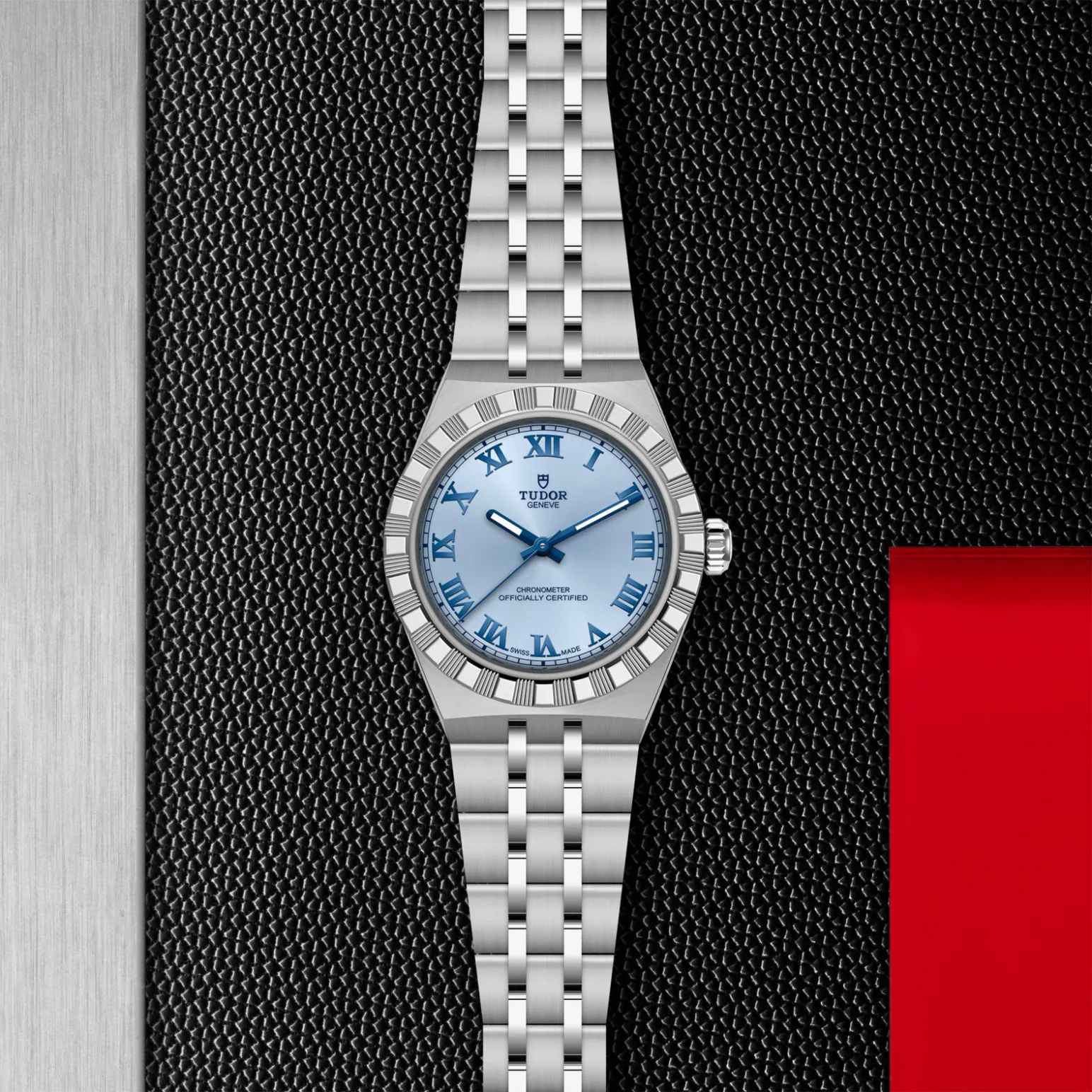 m2830a1a0-0004in_store_flat_lay | Brela Silver Tudor wristwatch with a light blue dial and Roman numeral markers resting on a textured black surface with a hint of red panel nearby | Brela