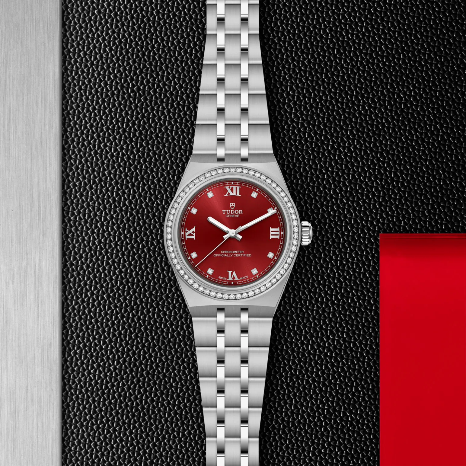 m2830a1s0-0002in_store_flat_lay | Brela Tudor Genève wristwatch with a red dial, diamond-bezel, and stainless steel bracelet on a black textured background. | Brela