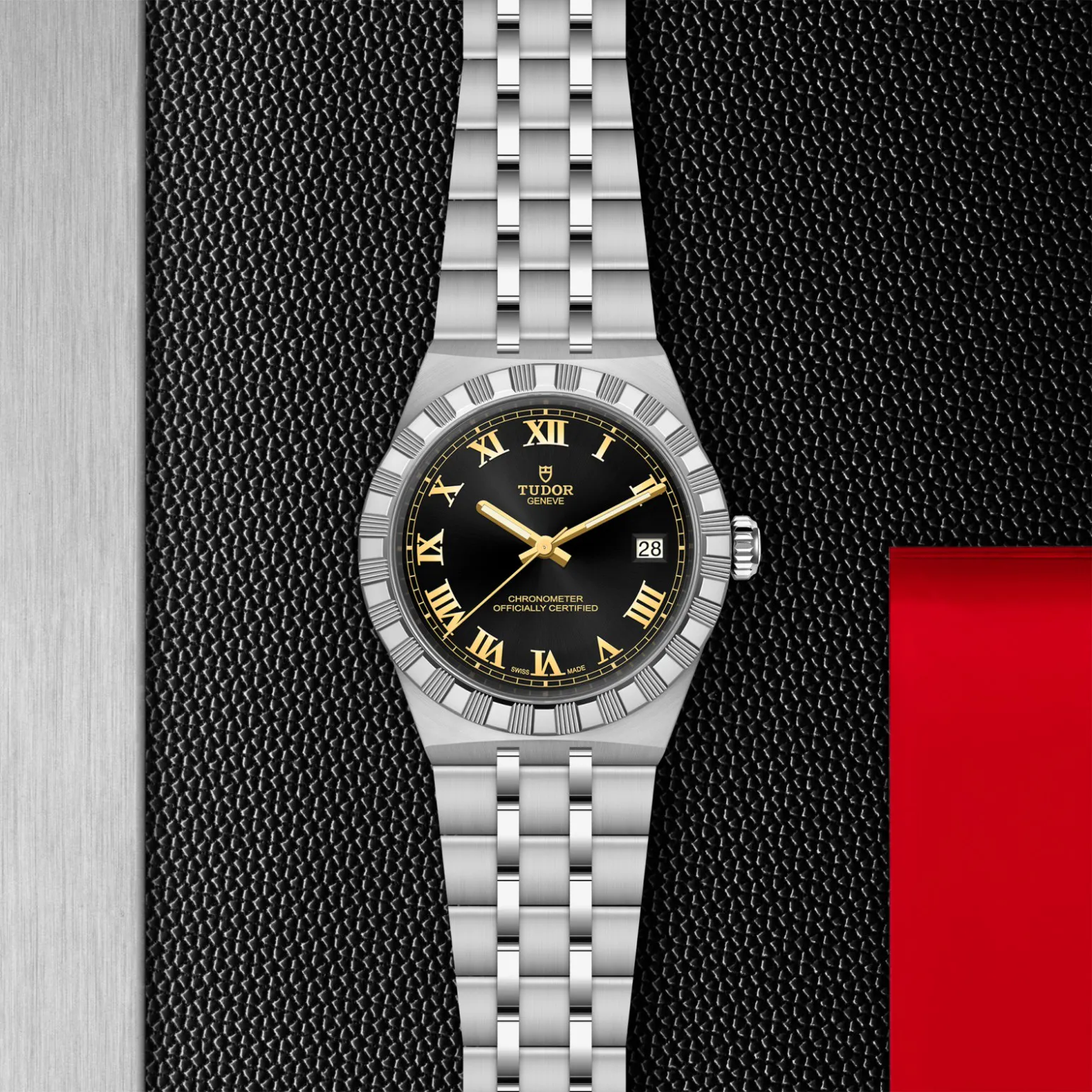 m2836c1a0-0101in_store_flat_lay | Brela Tudor wristwatch with black dial, gold Roman numerals, date window, and silver bracelet on a textured black backdrop. | Brela