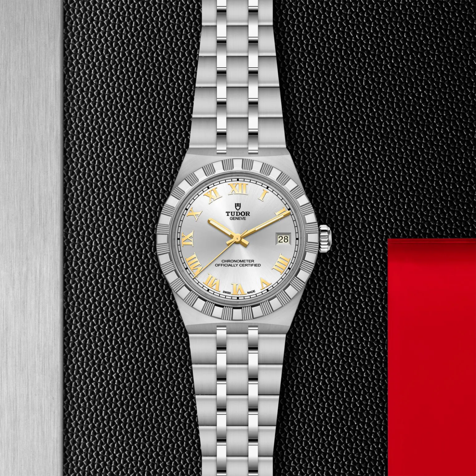 m2836c1a0-0104in_store_flat_lay | Brela Close-up of a silver Tudor wristwatch with gold Roman numerals and date window on a textured black background. | Brela