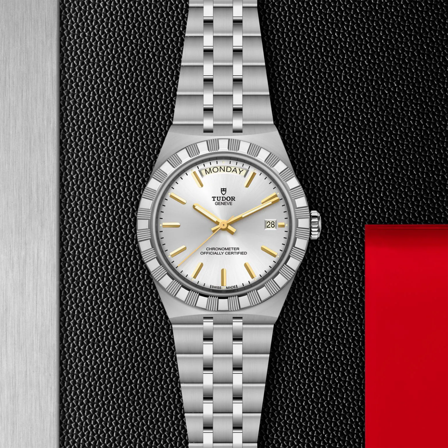 m2840d1a0-0004in_store_flat_lay | Brela Tudor stainless steel wristwatch with silver dial, gold hands and markers, day display at 12 o'clock and date at 3 o'clock on a black textured backdrop. | Brela