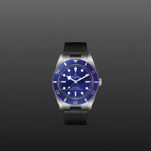 Close-up of a Tudor dive watch with a blue dial and blue bezel on a dark gradient background, black strap visible below the case.