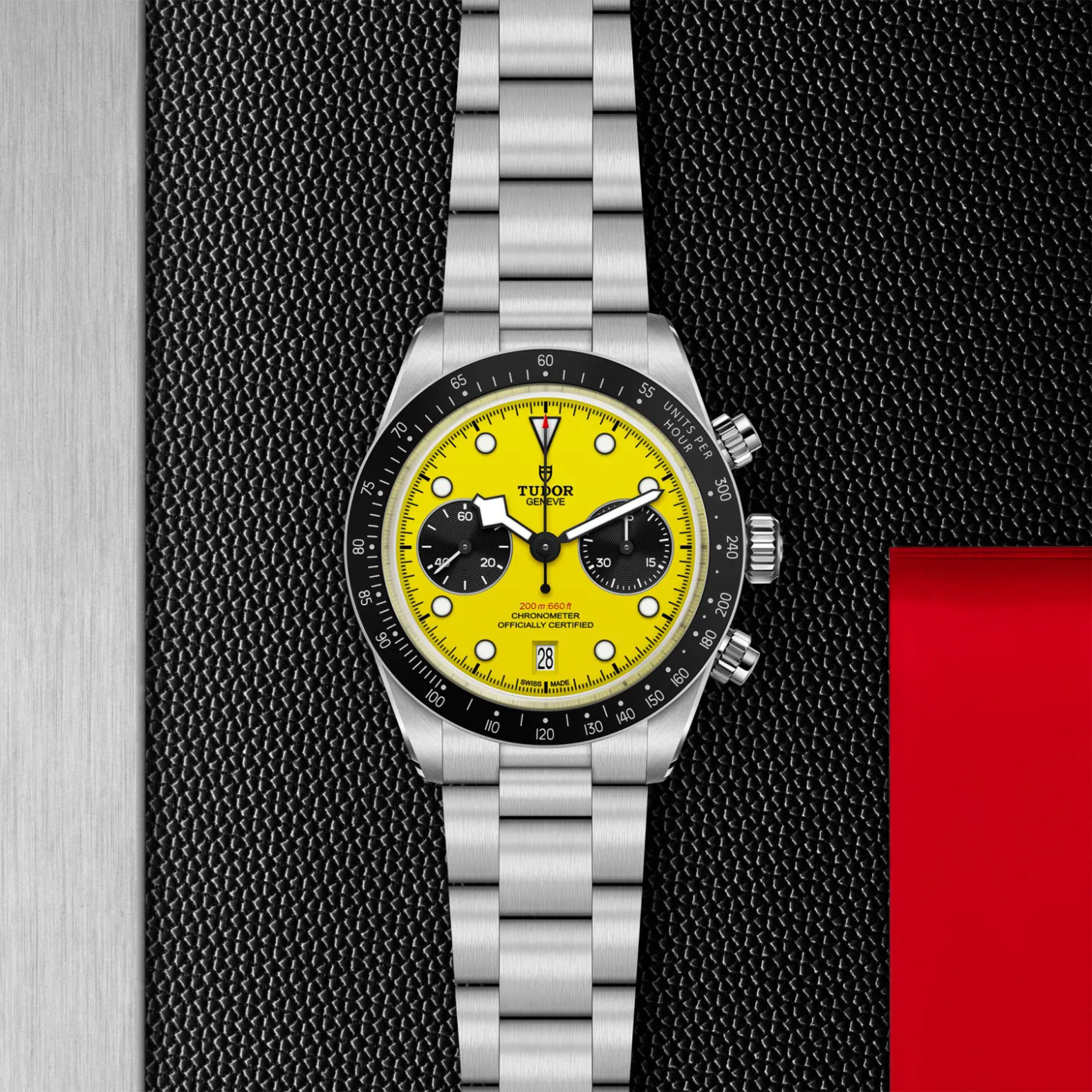 m79310n-0001in_store_flat_lay | Brela Tudor chronograph watch with a bright yellow dial and black subdials, stainless steel bracelet, on a textured black background with a silver strip on the left and a red panel on the right. | Brela