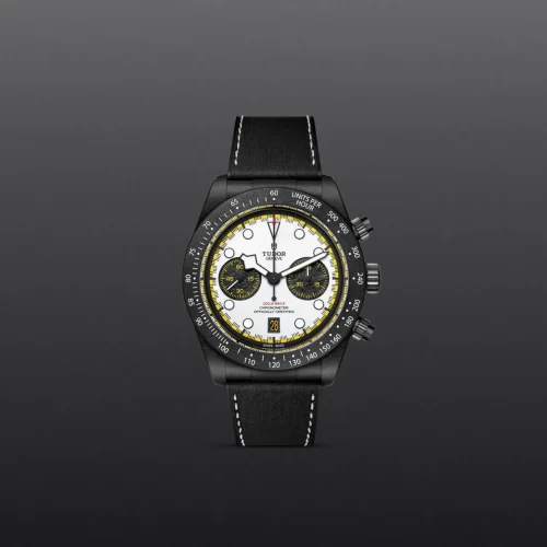 Tudor chronograph watch with white dial, black bezel, yellow accents, and black leather strap against a dark background.