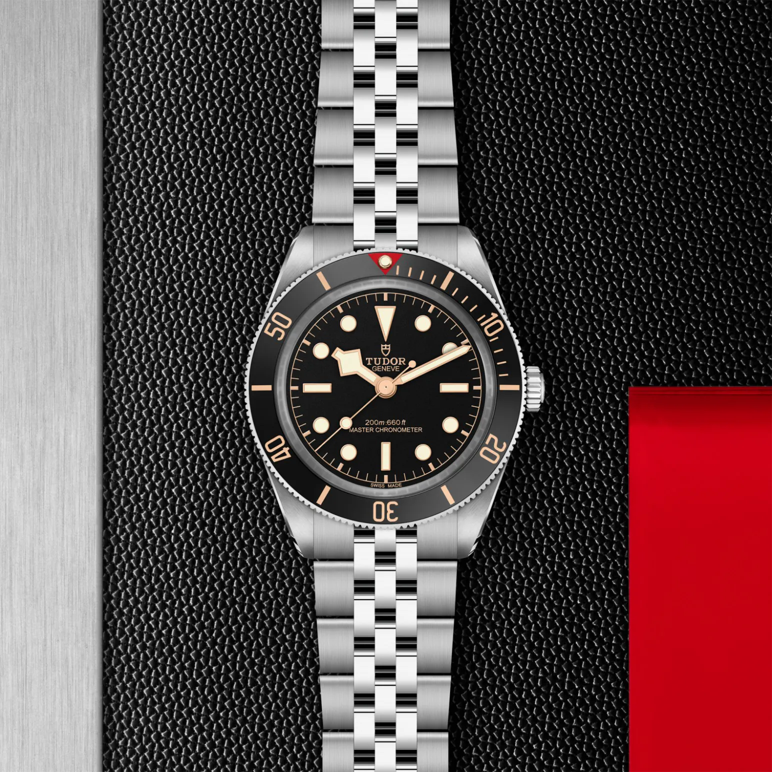 m7939a1a0nu-0001in_store_flat_lay | Brela Tudor wristwatch with black dial, gold hour markers, and stainless steel bracelet on a textured black surface with a red panel to the right. | Brela
