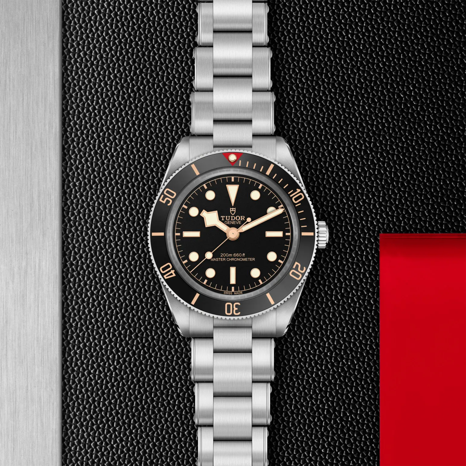m7939a1a0nu-0002in_store_flat_lay | Brela Tudor wristwatch with black dial and rose-gold markers on a stainless steel band, resting on a textured black surface with a red accent panel nearby | Brela