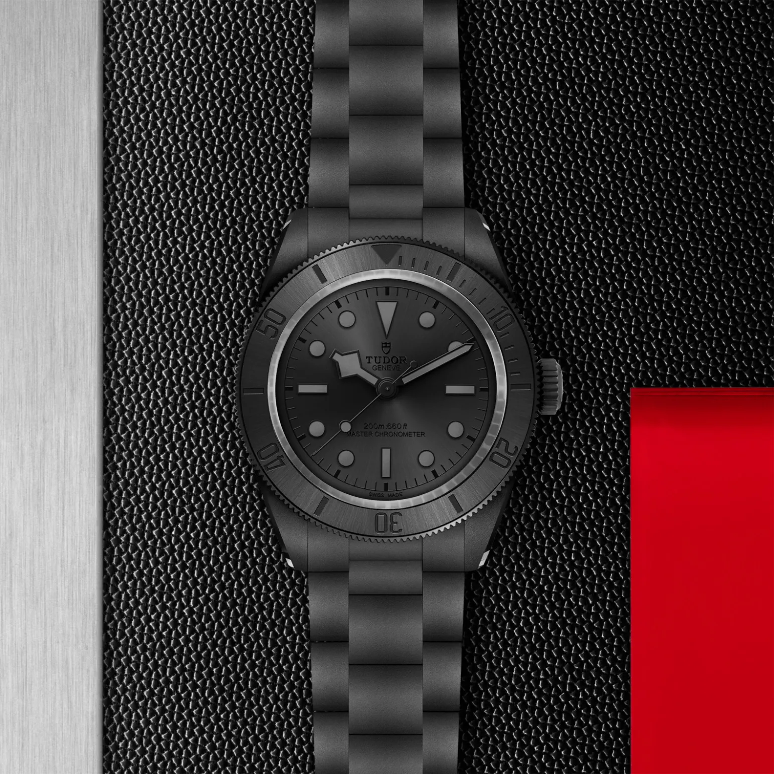 m7941a1acnu-0001in_store_flat_lay | Brela Close-up of a matte black Tudor diver's watch with a metal bracelet on a textured black surface, with a brushed silver strip on the left and a red block on the right. | Brela
