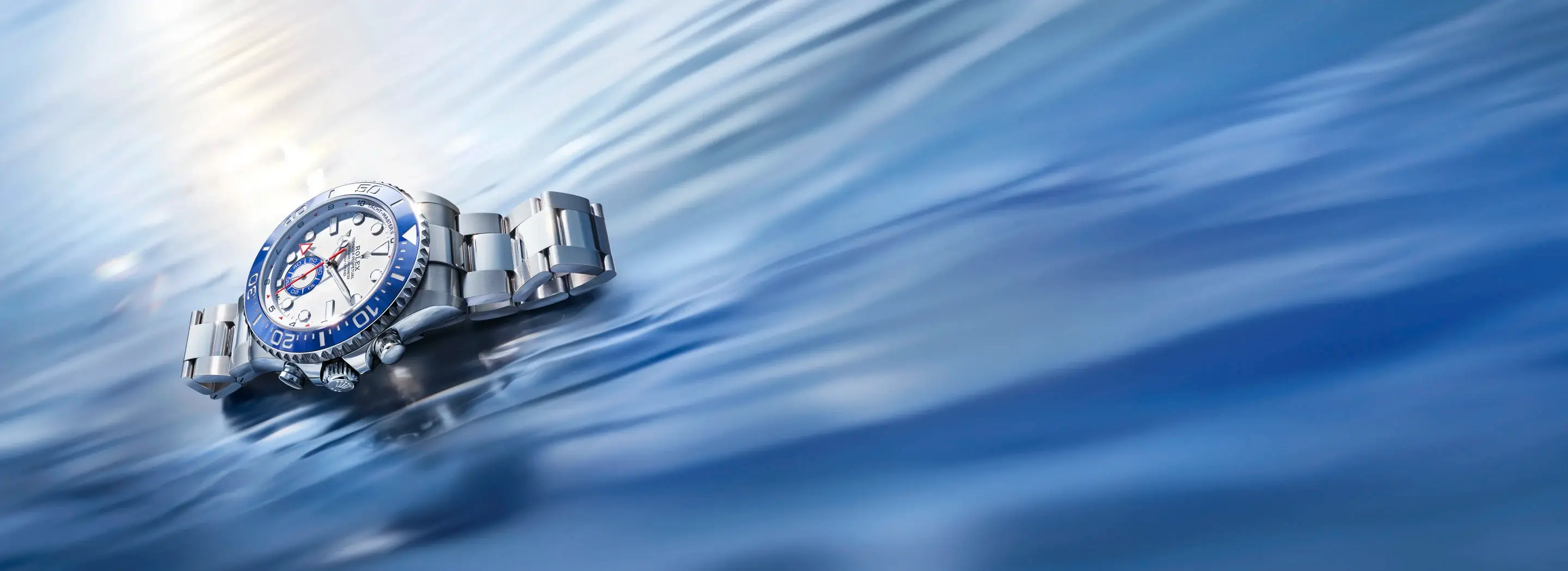 Silver stainless steel wristwatch with a blue bezel resting on a glossy, water-like blue surface.